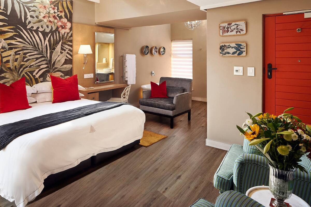 Une chambre d’hôtel avec un grand lit, un bureau, une chaise, une porte rouge et un vase rempli de fleurs.