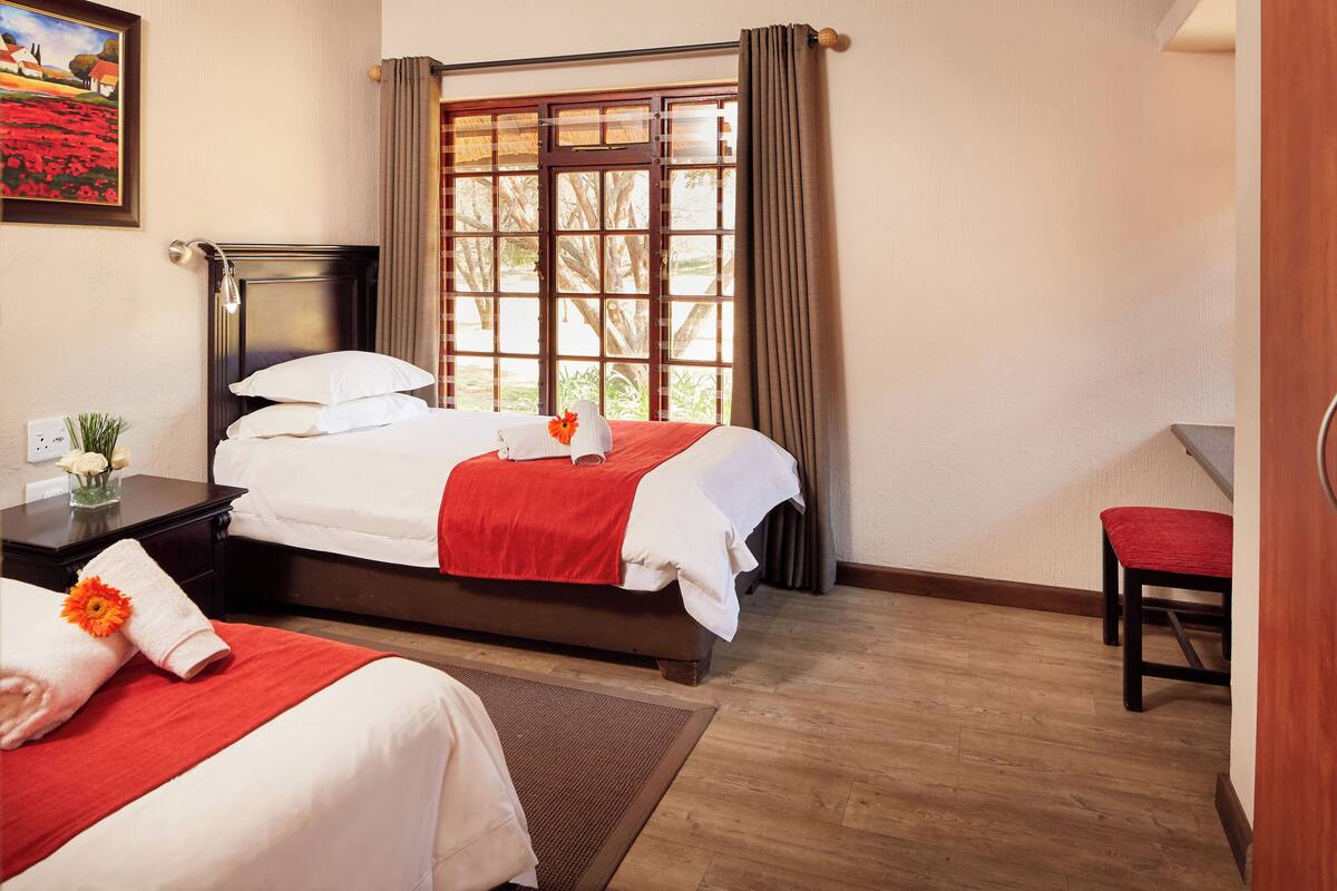 Une chambre d’hôtel avec deux lits, un plancher en bois, une fenêtre avec des rideaux, un bureau avec une lampe et un tableau au mur.