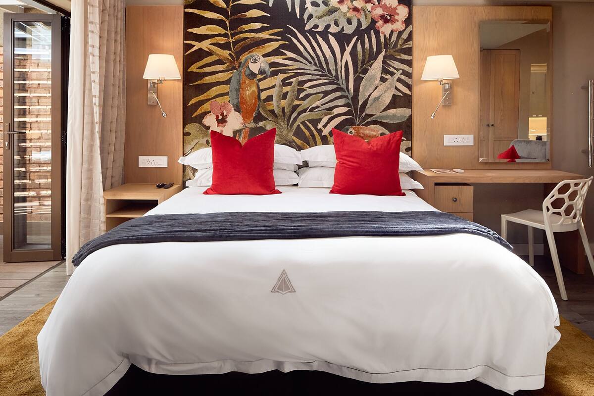 Une chambre d’hôtel avec un grand lit, deux oreillers rouges, une table de chevet, un bureau, une chaise et un grand miroir.