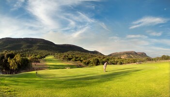 Un campo de golf con una persona vestida con camisa blanca y pantalones cortos, cielo despejado y montañas al fondo.