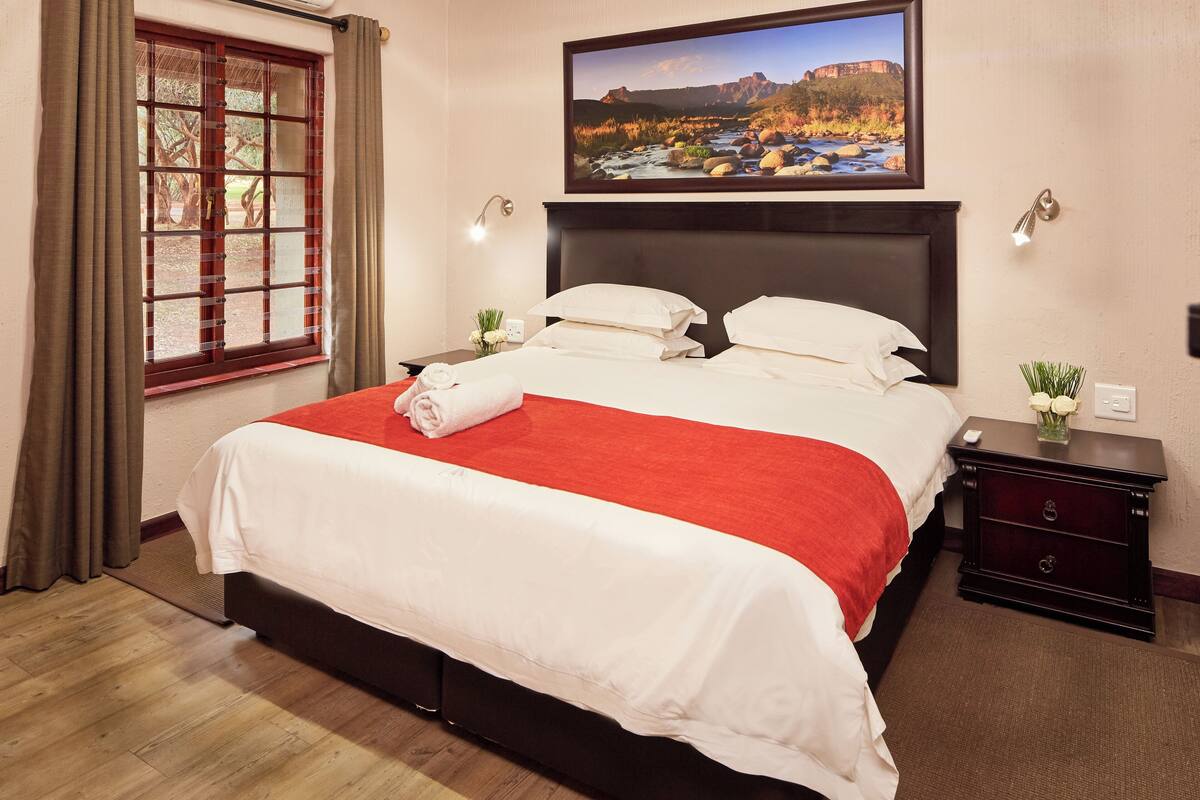 Une chambre d’hôtel avec un grand lit, une table de chevet ornée d’un vase de fleurs, un tableau au mur et une fenêtre avec des rideaux.