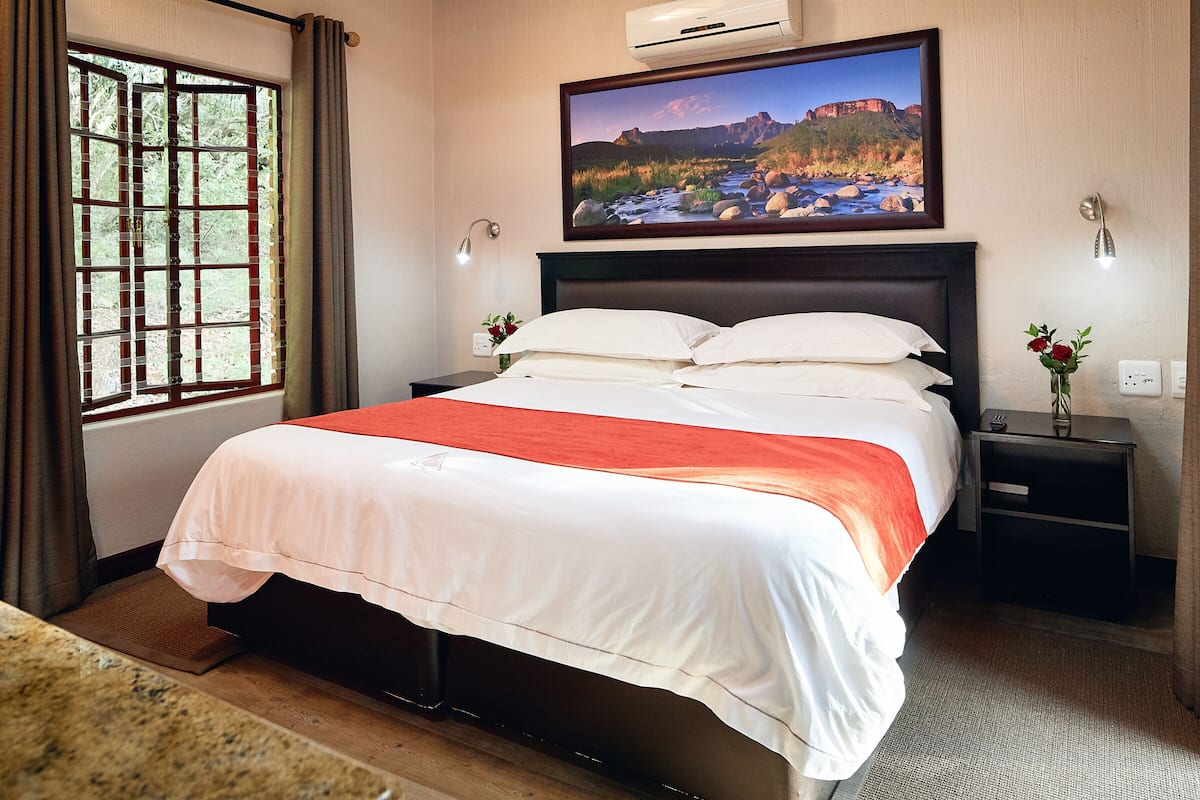 Une chambre d’hôtel avec un grand lit, une fenêtre avec des rideaux, un tableau au mur et une table de chevet ornée d’un vase de fleurs.