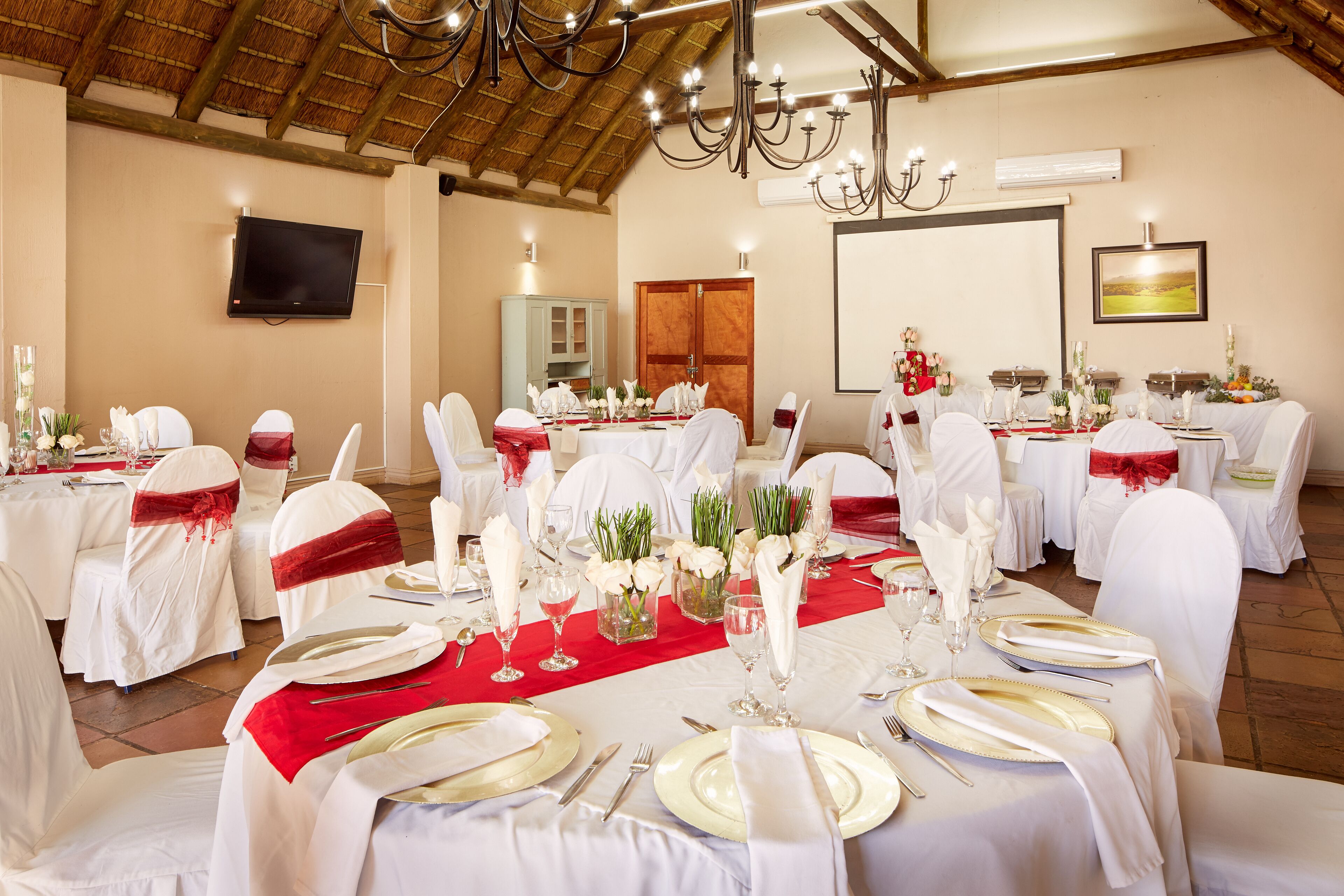 A banquet hall with round tables set for a formal event, featuring white tablecloths, gold cutlery, and red sashes on chairs.