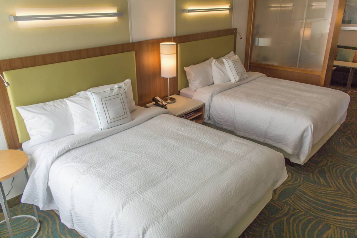 A hotel room with two beds, a nightstand with a lamp, and a small table.