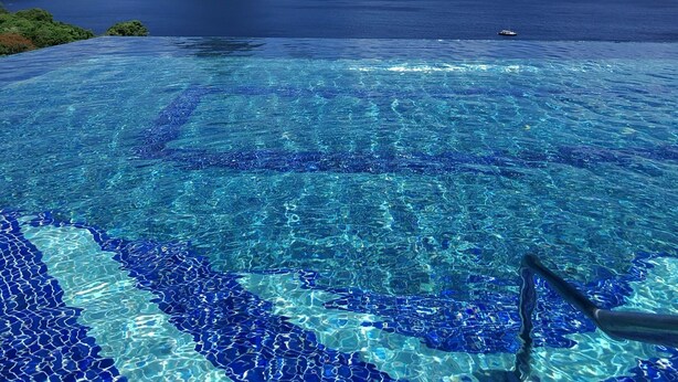Une piscine d’un bleu éclatant, ornée de motifs en mosaïque, entourée d’une végétation luxuriante et offrant une vue sur une côte montagneuse sous un ciel d’un bleu profond.