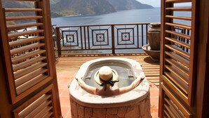 Une terrasse en bois avec un jacuzzi en pierre et une vue sur la montagne et la mer.