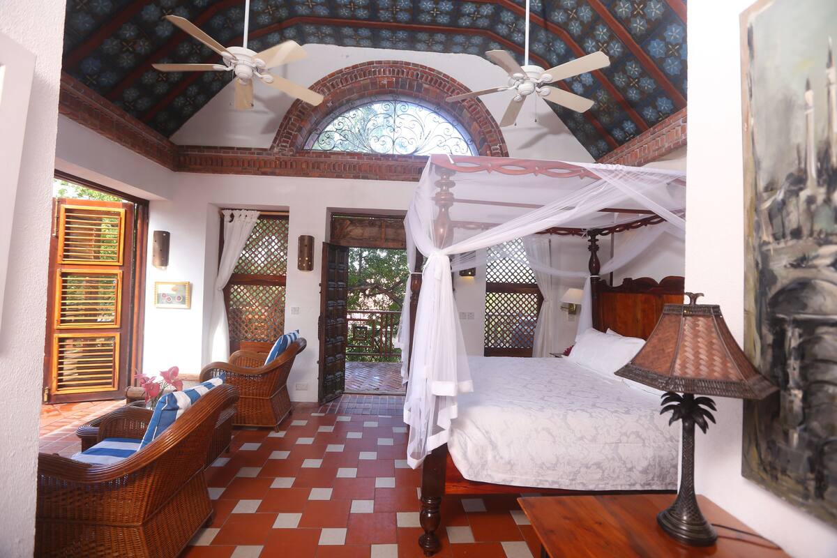 Une pièce comprenant un lit, deux fauteuils en osier, une table, un ventilateur de plafond et une lampe.
