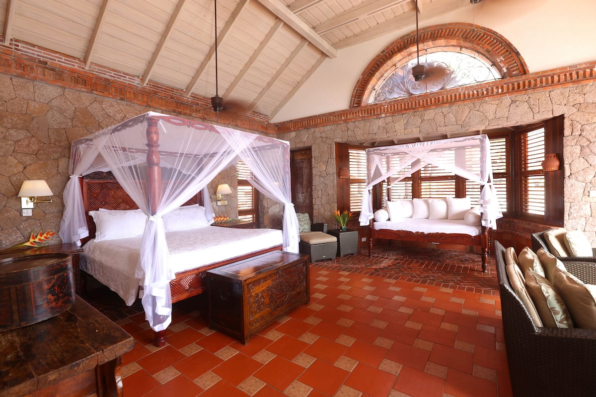 A room with a large bed, a smaller bed, a wooden bench, and a wicker sofa.