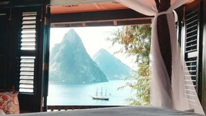 Une chambre avec un lit, une fenêtre ornée de rideaux blancs et une vue sur des montagnes et une étendue d’eau.