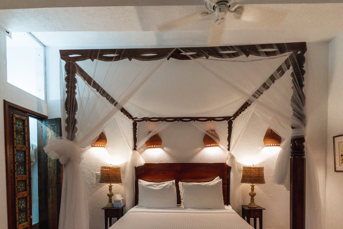 A four-poster bed with a canopy, two bedside tables with lamps, and a checkered floor pattern.