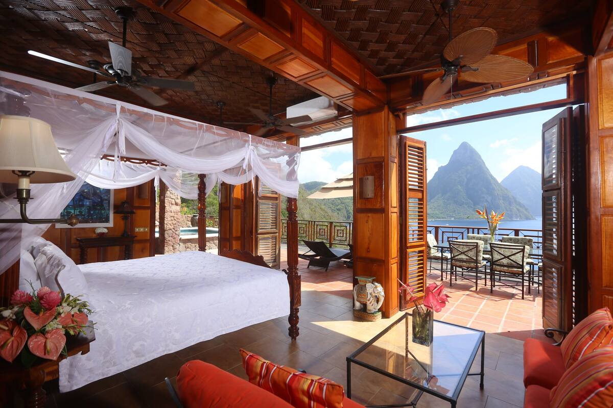 A bedroom with a bed, a sitting area with a table and chairs, and a balcony with a view of a mountain and the sea.