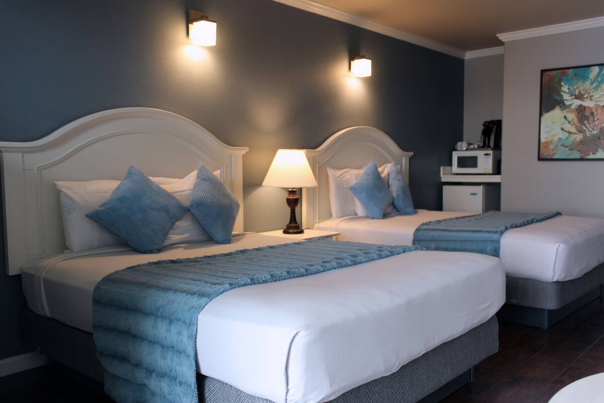 A hotel room with two beds, each with a white headboard, blue bedding, and a lamp on the nightstand.