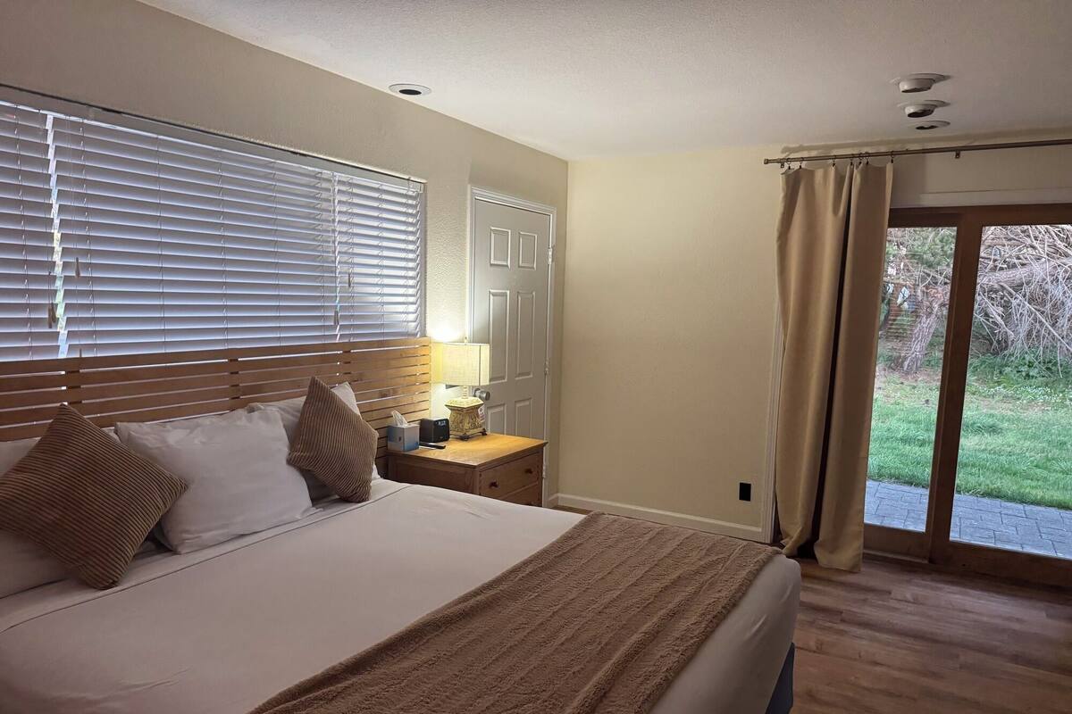 A bedroom with a large bed, wooden headboard, bedside table with lamp, and a view of a garden through a sliding glass door.