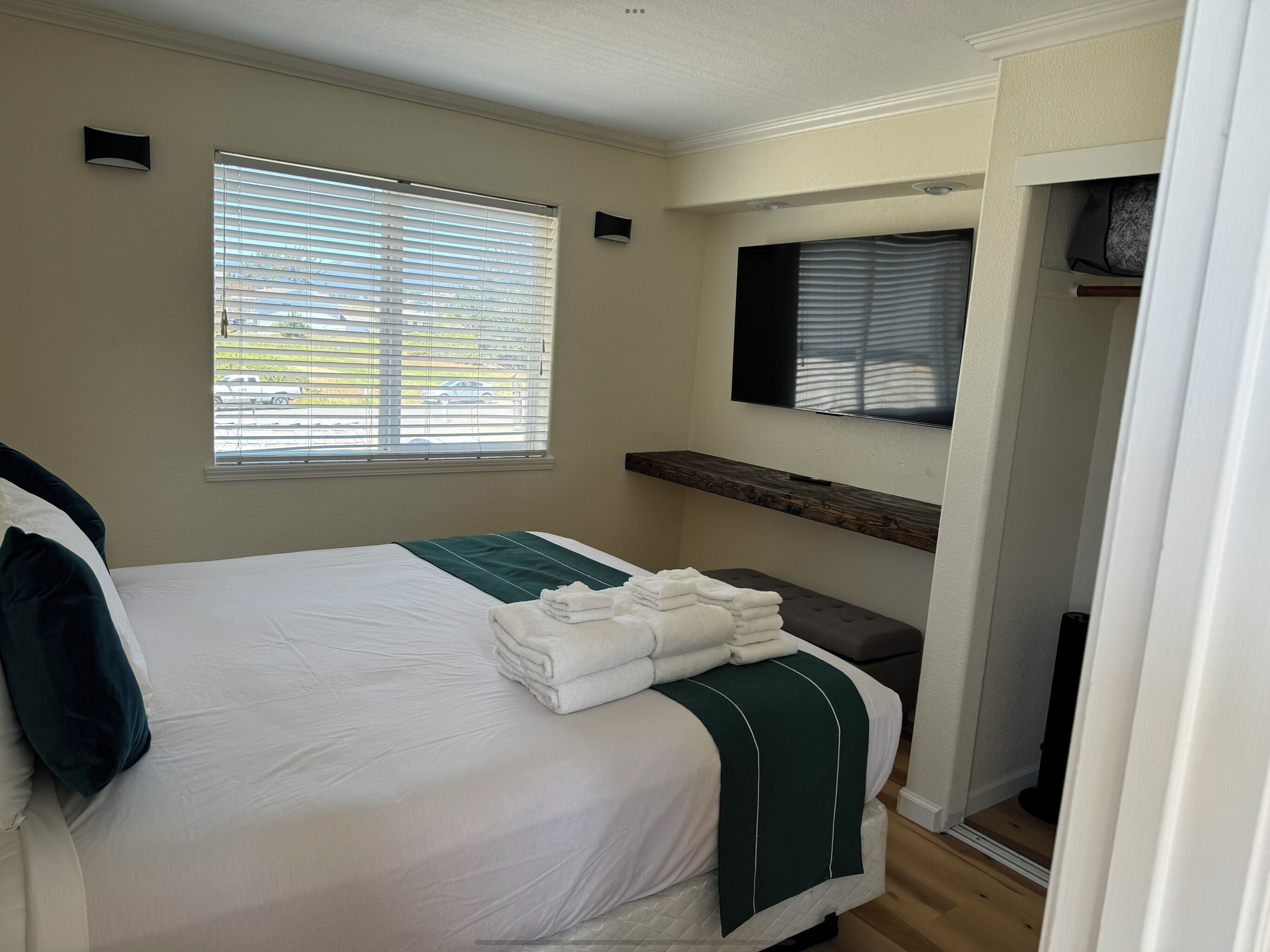 A hotel room with a bed, a window with blinds, a mounted TV, and a small shelf.