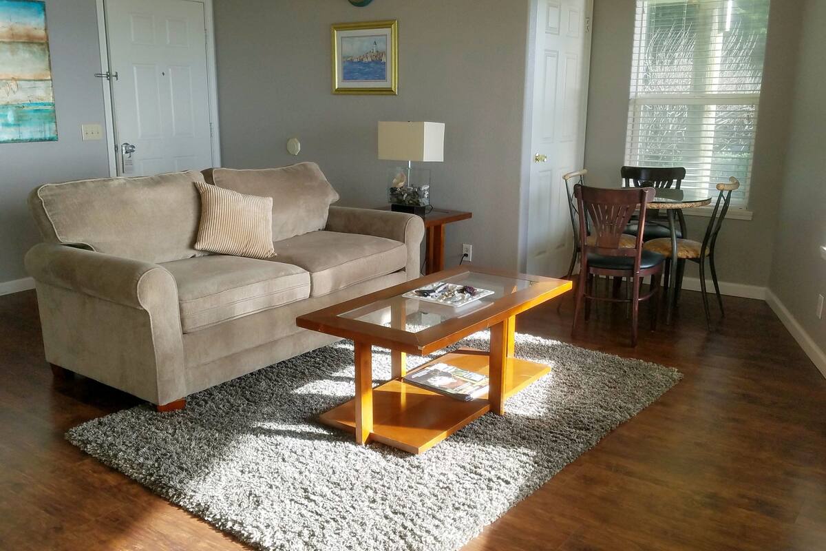 A living room with a beige sofa, a wooden coffee table, a dining table with chairs, a lamp, and a painting on the wall.