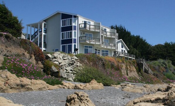 A multi-story coastal building with balconies, situated on a rocky hillside with vegetation and flowers.