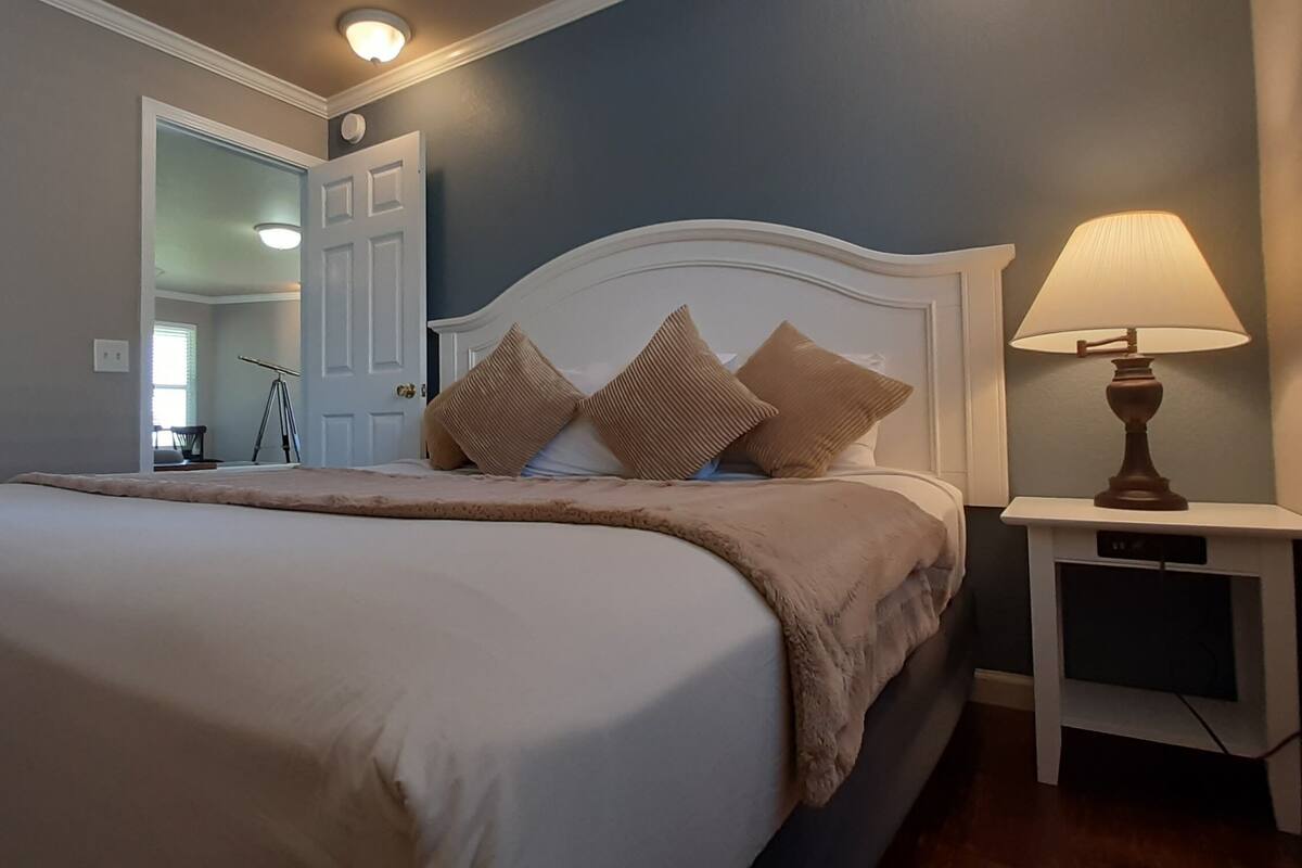 A neatly made bed with a white comforter and brown throw, a bedside lamp, and a small bedside table with a TV.