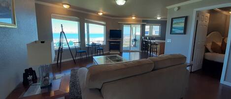 A spacious living room with a large sofa, a coffee table, and a TV. There are multiple windows offering a view of the ocean.