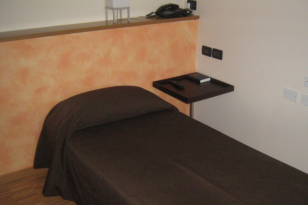 Una cama individual con cubrecolor oscuro en una habitación con piso de madera y una mesita con teléfono y control remoto.