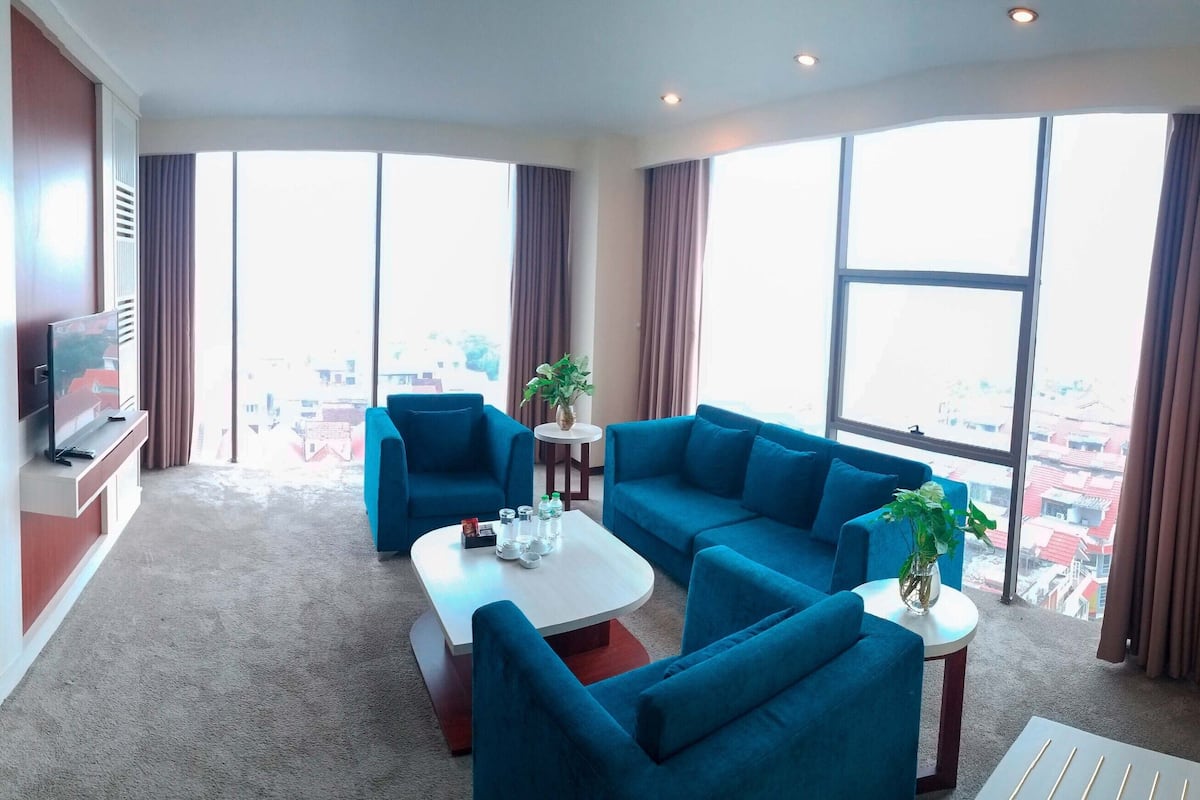 A hotel room with a bed, a TV, a sofa set, a coffee table, and large windows offering a city view.