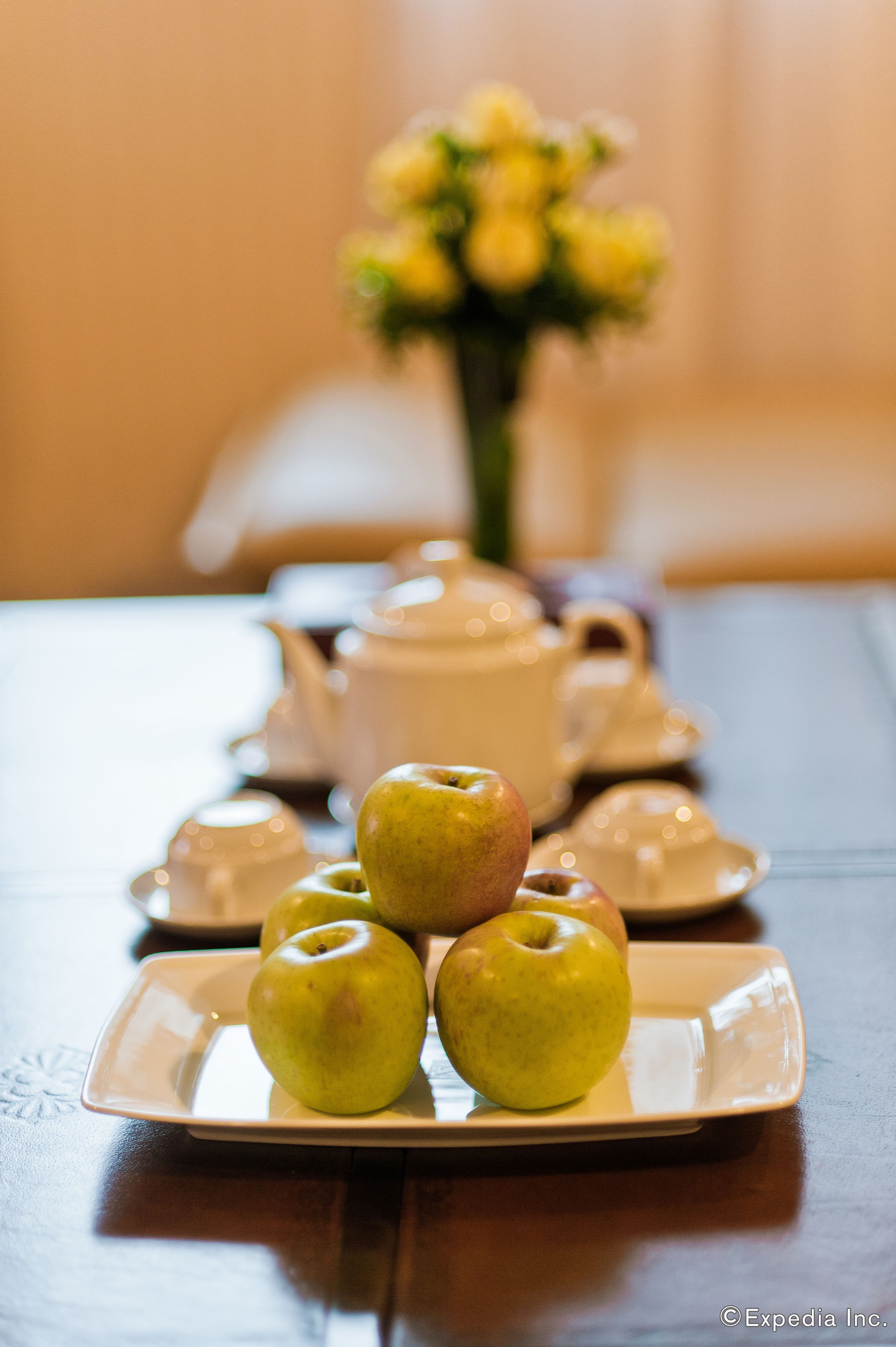 A table with a plate of apples, a teapot, cups, and a vase with flowers.