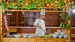 A chef in a kitchen with a wooden counter, various cooking utensils, and a decorative hanging with fruits and leaves. - Muong Thanh Dien Chau Hotel (Dien Chau)