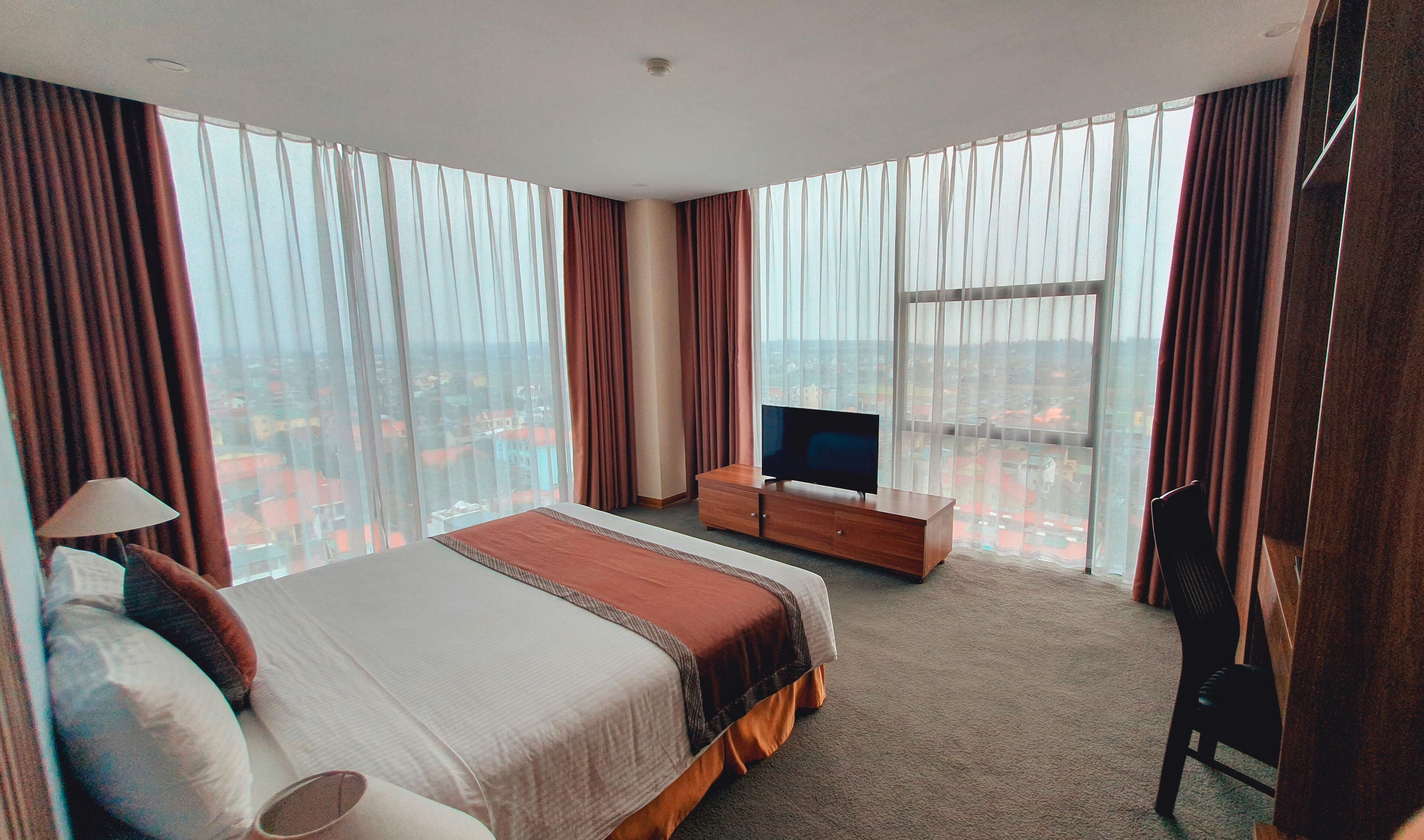 A hotel room with a large bed, a TV on a wooden cabinet, and a view of a cityscape through floor-to-ceiling windows.