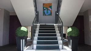 Un escalier moderne, surmonté d’un tableau aux motifs géométriques, et flanqué de deux sphères de topiaire disposées dans des pots.