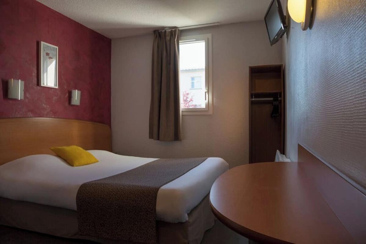 Ein Hotelzimmer mit Bett, Nachttisch, einem Fenster mit Vorhängen, einem an der Wand montierten Fernseher und einem Kleiderschrank.