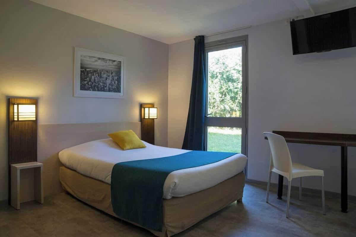 Ein Hotelzimmer mit Bett, Nachttischlampen, Schreibtisch, Stuhl, Fernseher und einem Fenster mit Blick auf Grünflächen.