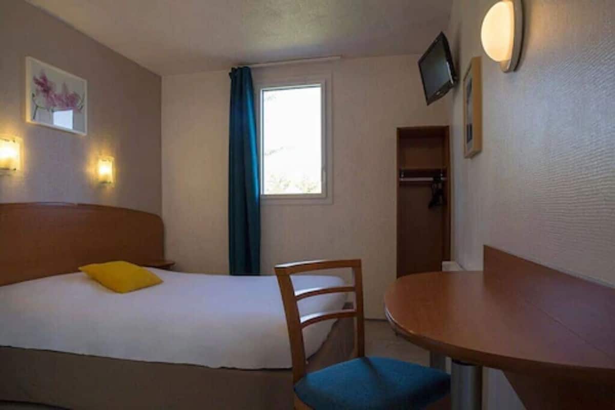 Ein Hotelzimmer mit einem Bett, einem Sessel, einem kleinen Tisch, einem Fenster mit Vorhängen und Wandleuchten.