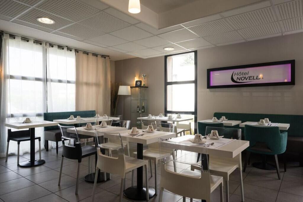 A hotel dining area with tables and chairs, a wall-mounted TV displaying the hotel name, and a window with curtains.