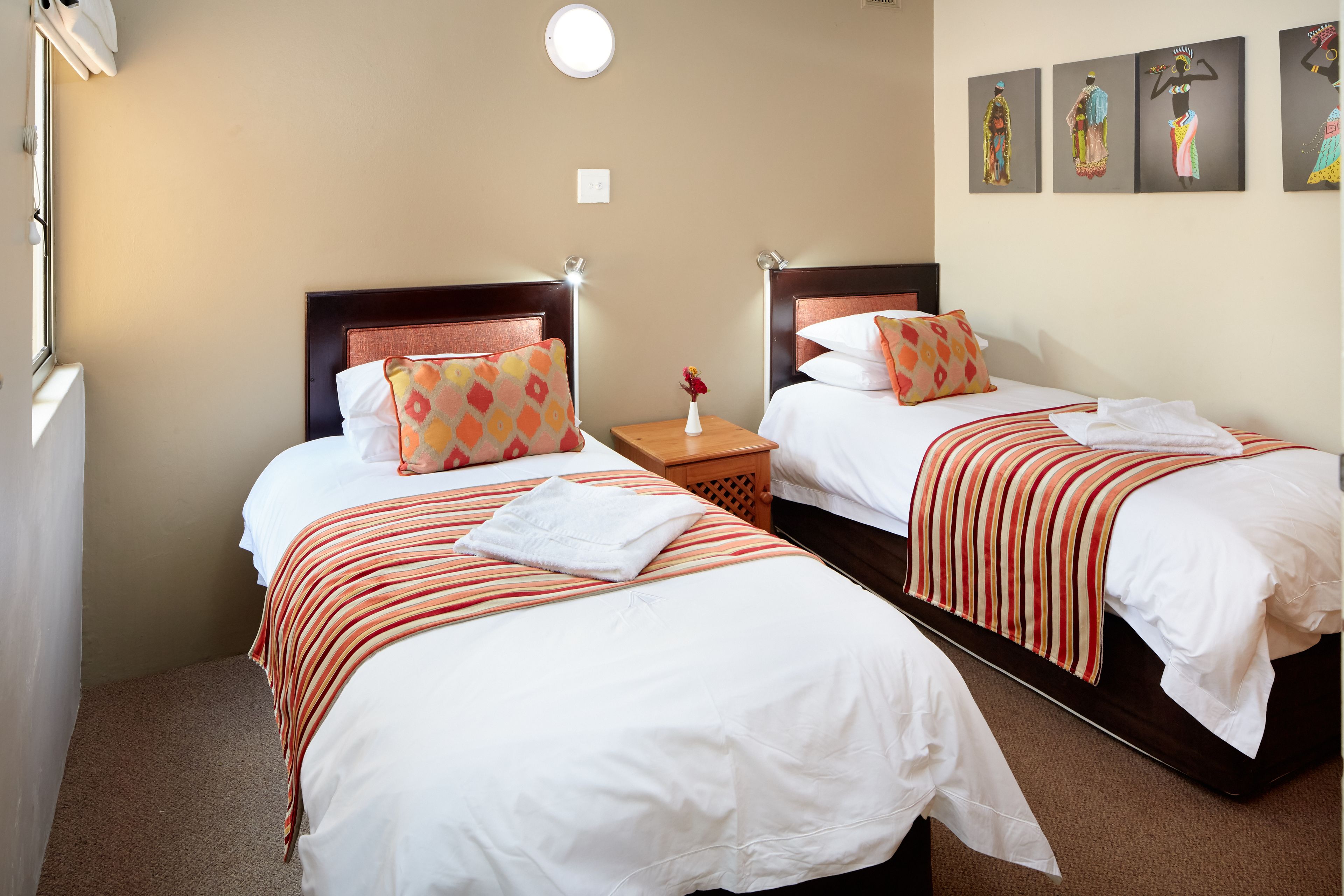 A hotel room with two beds, each with a wooden headboard, white linens, and striped blankets. There is a nightstand with a vase of flowers and a lamp between the beds.