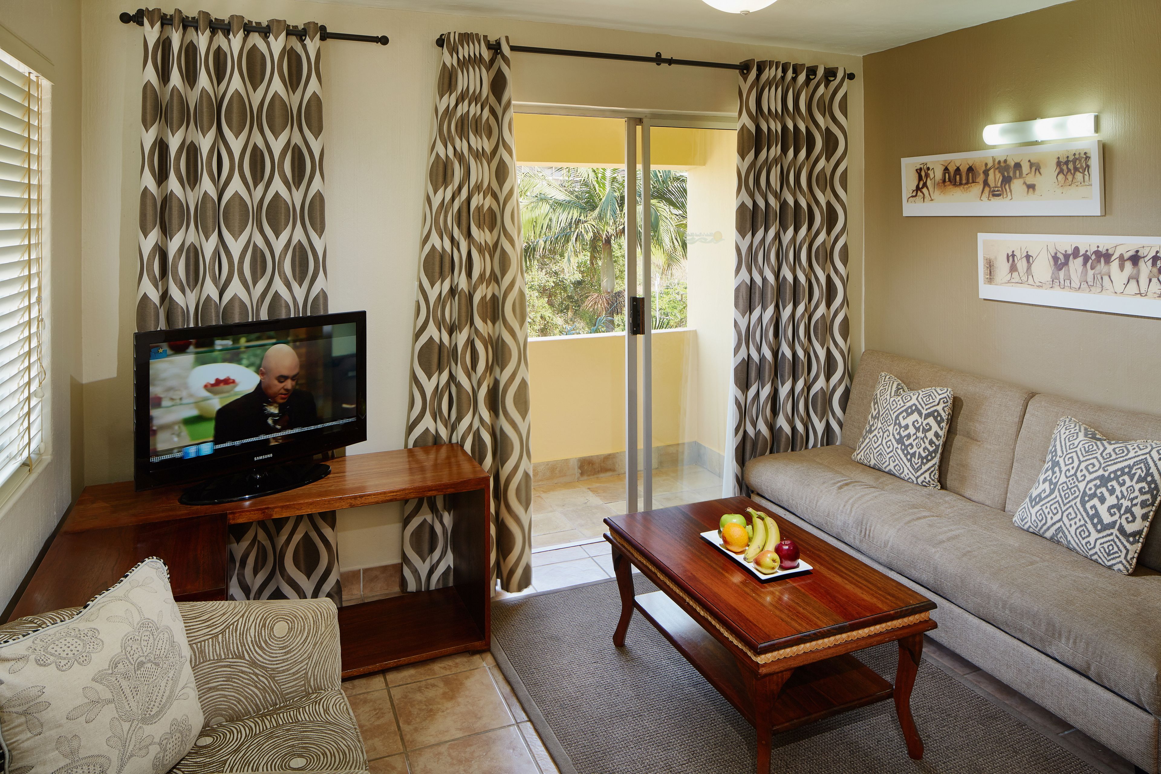 A living room with a TV, a wooden coffee table with fruit, a sofa, patterned curtains, and a balcony with a view.