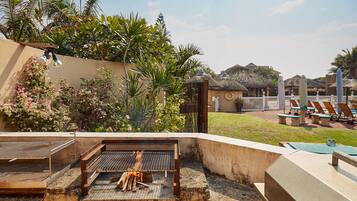 An outdoor grill area with a thatched-roof structure, surrounded by lush greenery and a poolside lounge area with sun loungers.