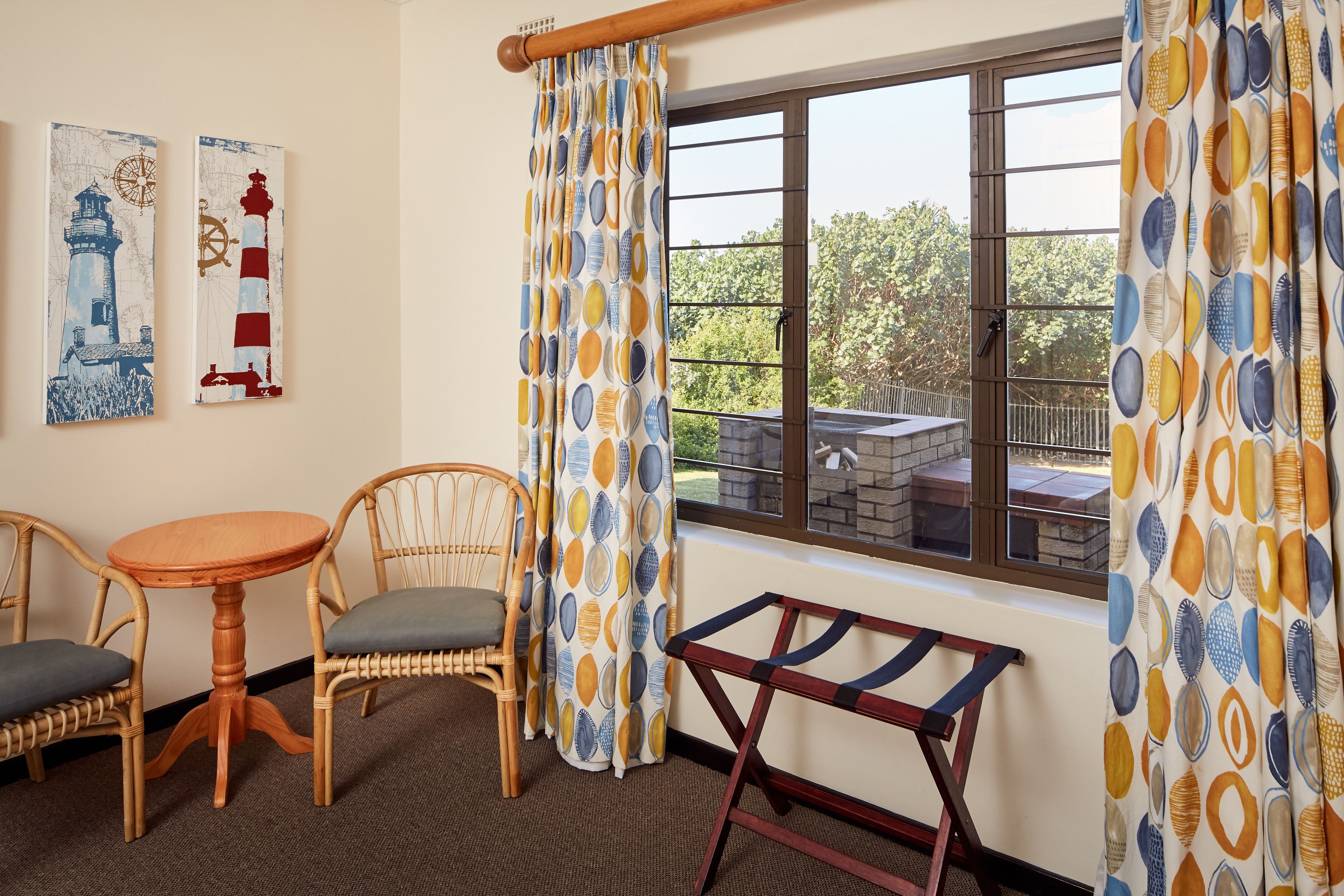 A room with a wooden table and two wicker chairs, a window with curtains, and two wall-mounted paintings.