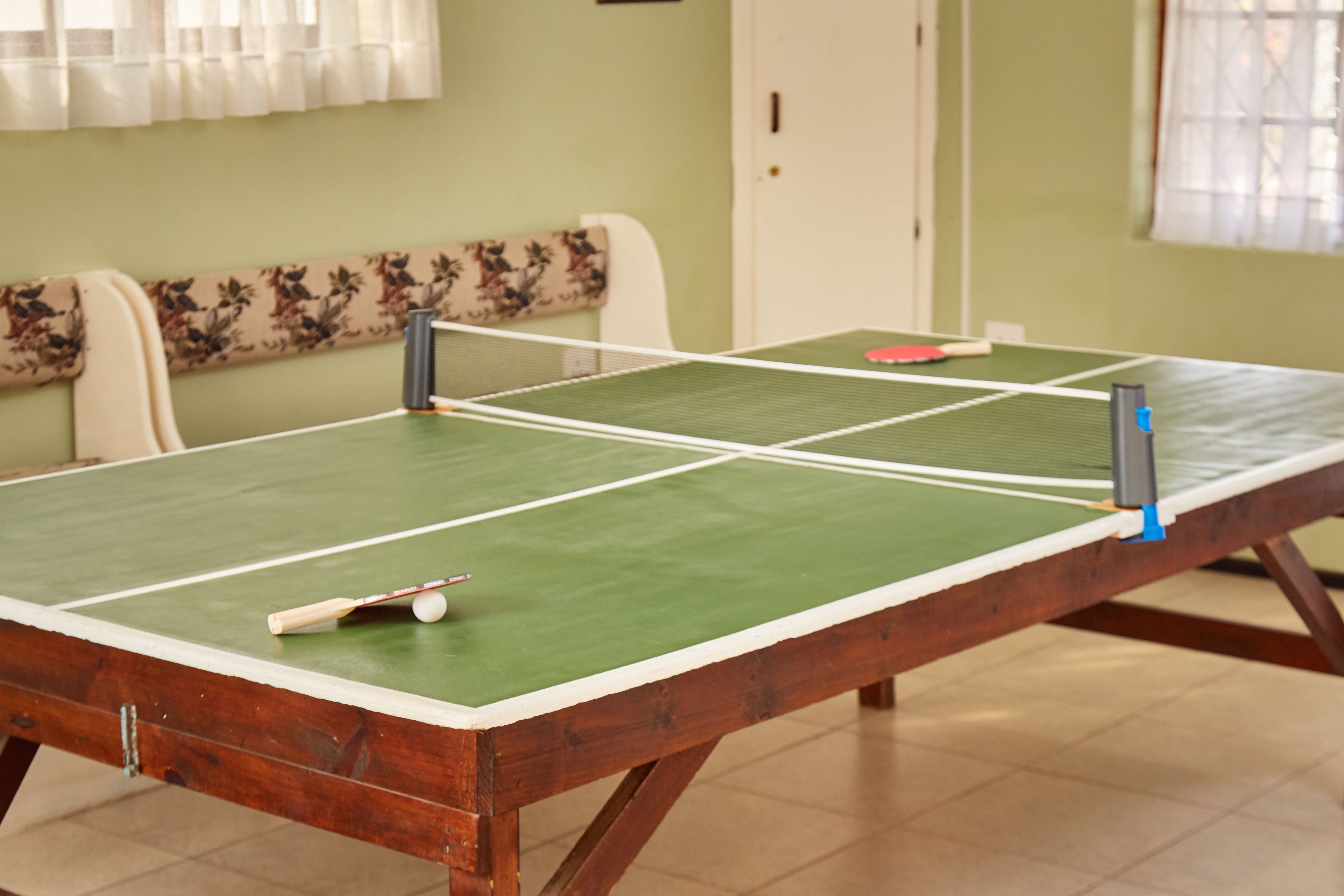 A ping pong table with a net, a paddle, and a ball in a room with a floral bench and a door in the background.