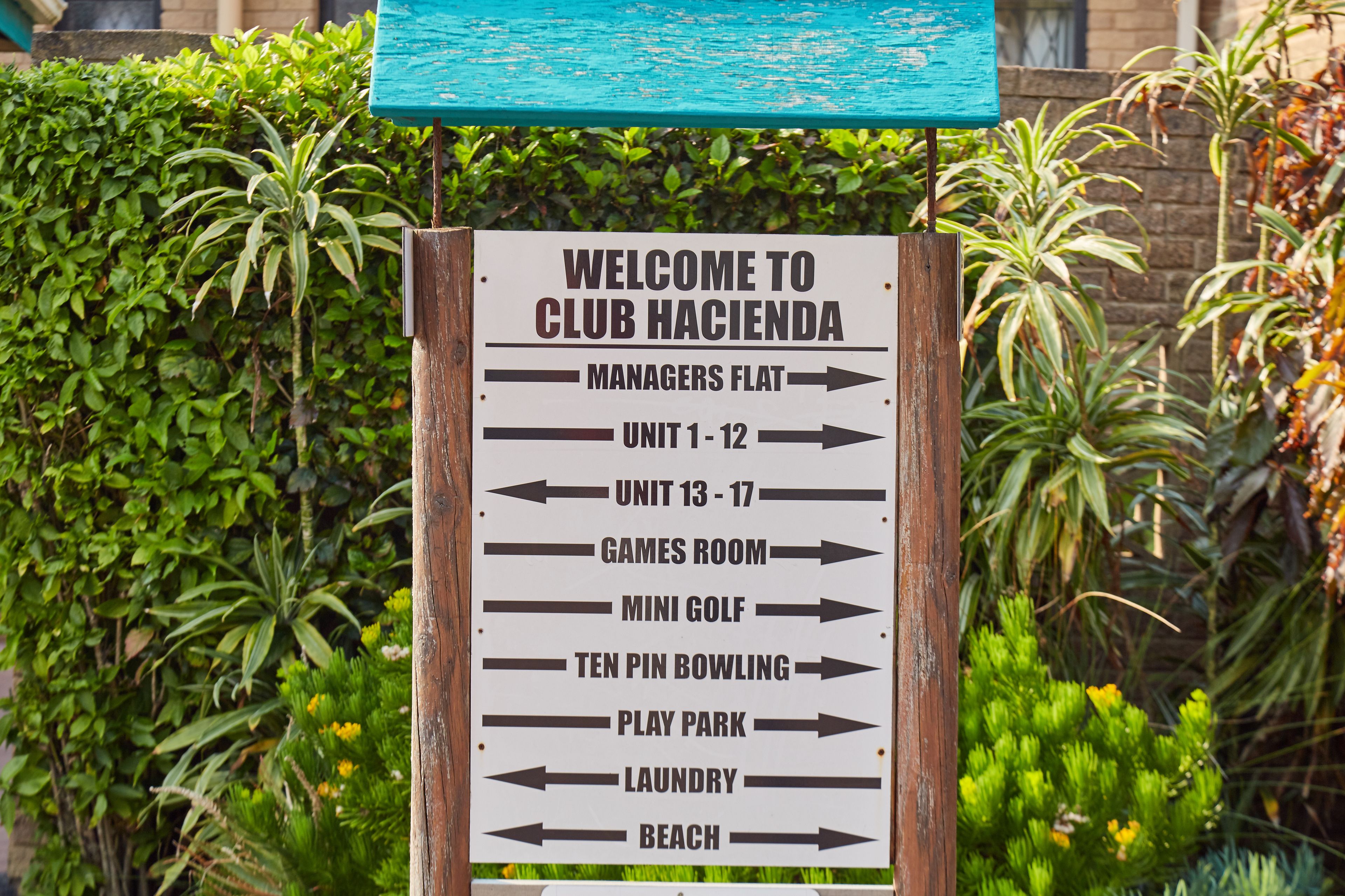 Hinweisschild für Club Hacienda mit Pfeilen zu verschiedenen Annehmlichkeiten wie Managerwohnung, Einheiten 1-12, Spielraum, Minigolf, Kegelbahn, Spielplatz, Wäscherei und Strand.