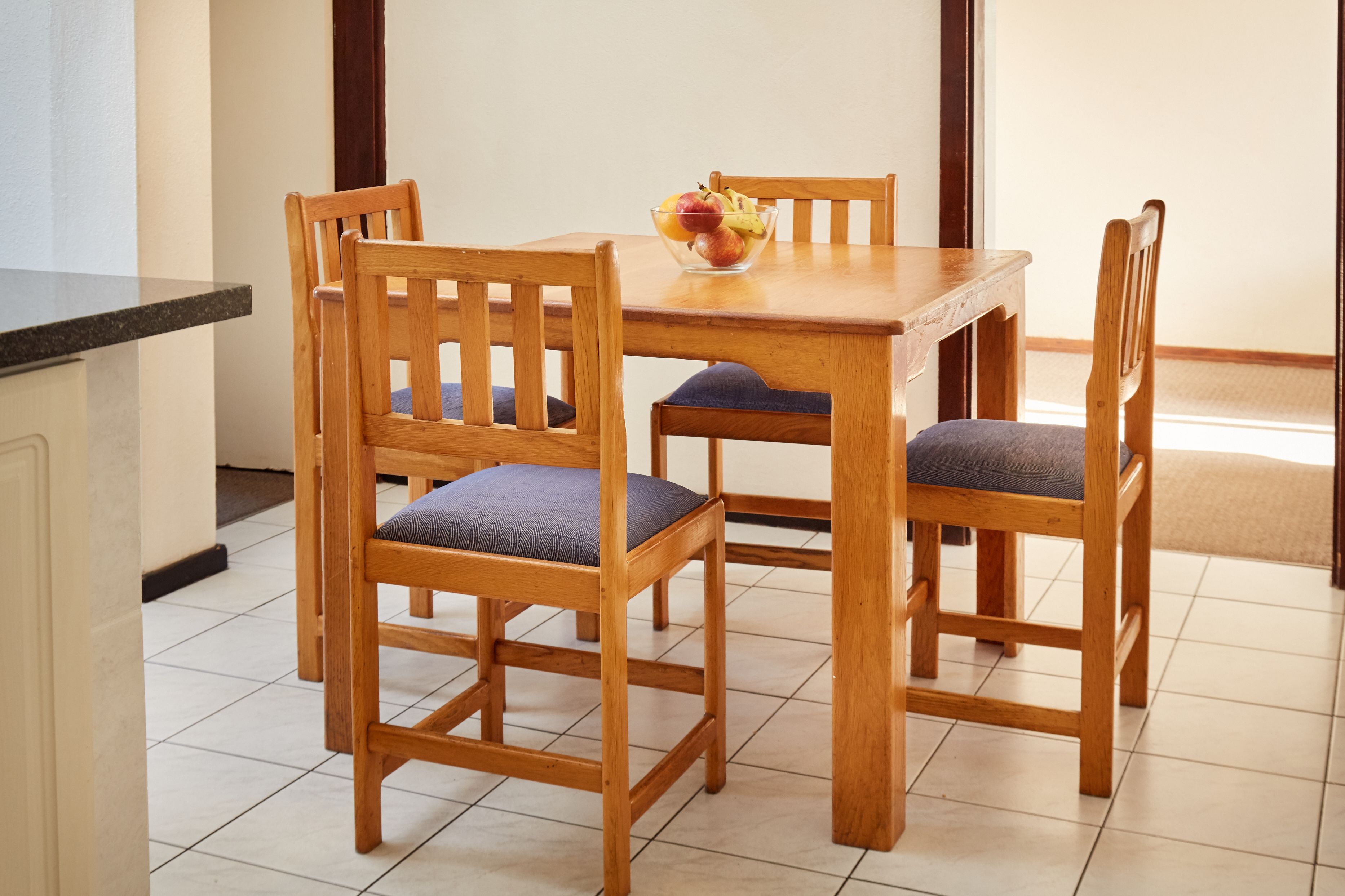 A wooden dining table with four chairs, a bowl of fruit, and a tiled floor.