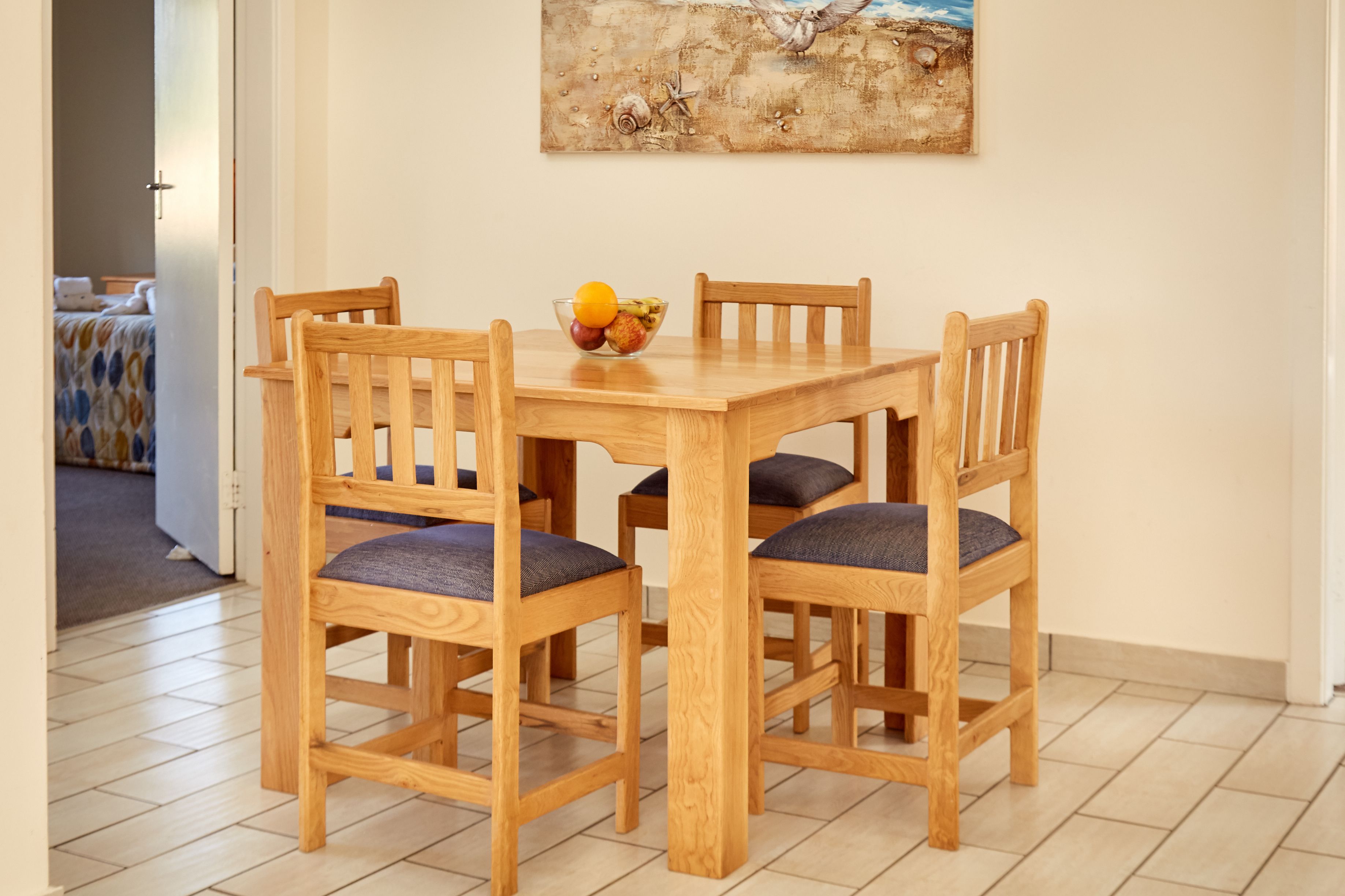 Een houten eettafel met vier stoelen, een schaal fruit en een schilderij aan de muur.