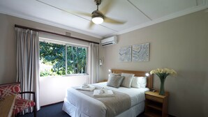 A hotel room with a large bed, a desk, a chair, a nightstand with a vase of flowers, and a ceiling fan. - First Group Lake View Cabanas (St. Lucia)