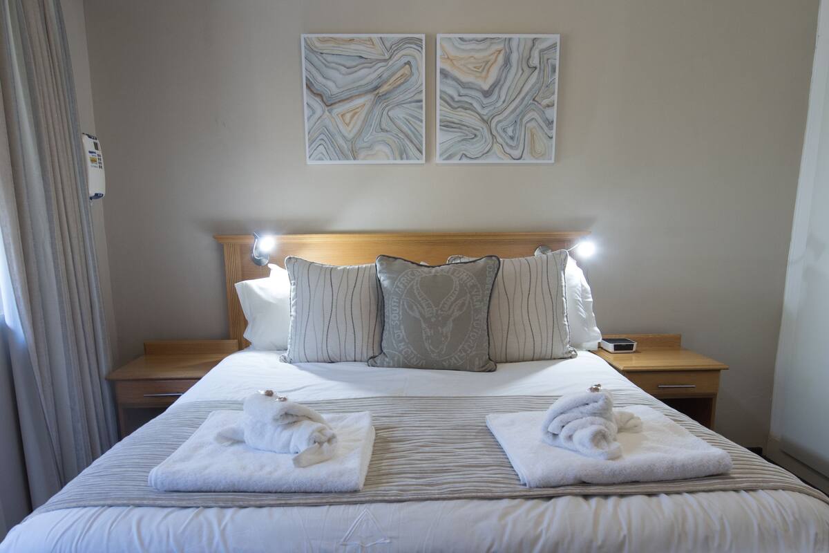 Ett hotellrum med en stor säng, två kuddar, ett sängbord med en lampa och två konstverk på väggen.
