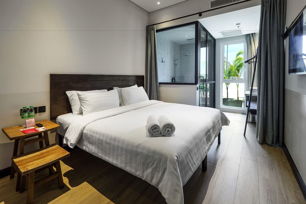 A hotel room with a large bed, a wooden nightstand, a small table, and a bench. There is a window with curtains and a view of greenery outside.