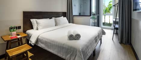 Een hotelkamer met een groot bed, een houten nachtkastje, een klein tafeltje en een bankje. Er is een raam met gordijnen en uitzicht op groen buiten.