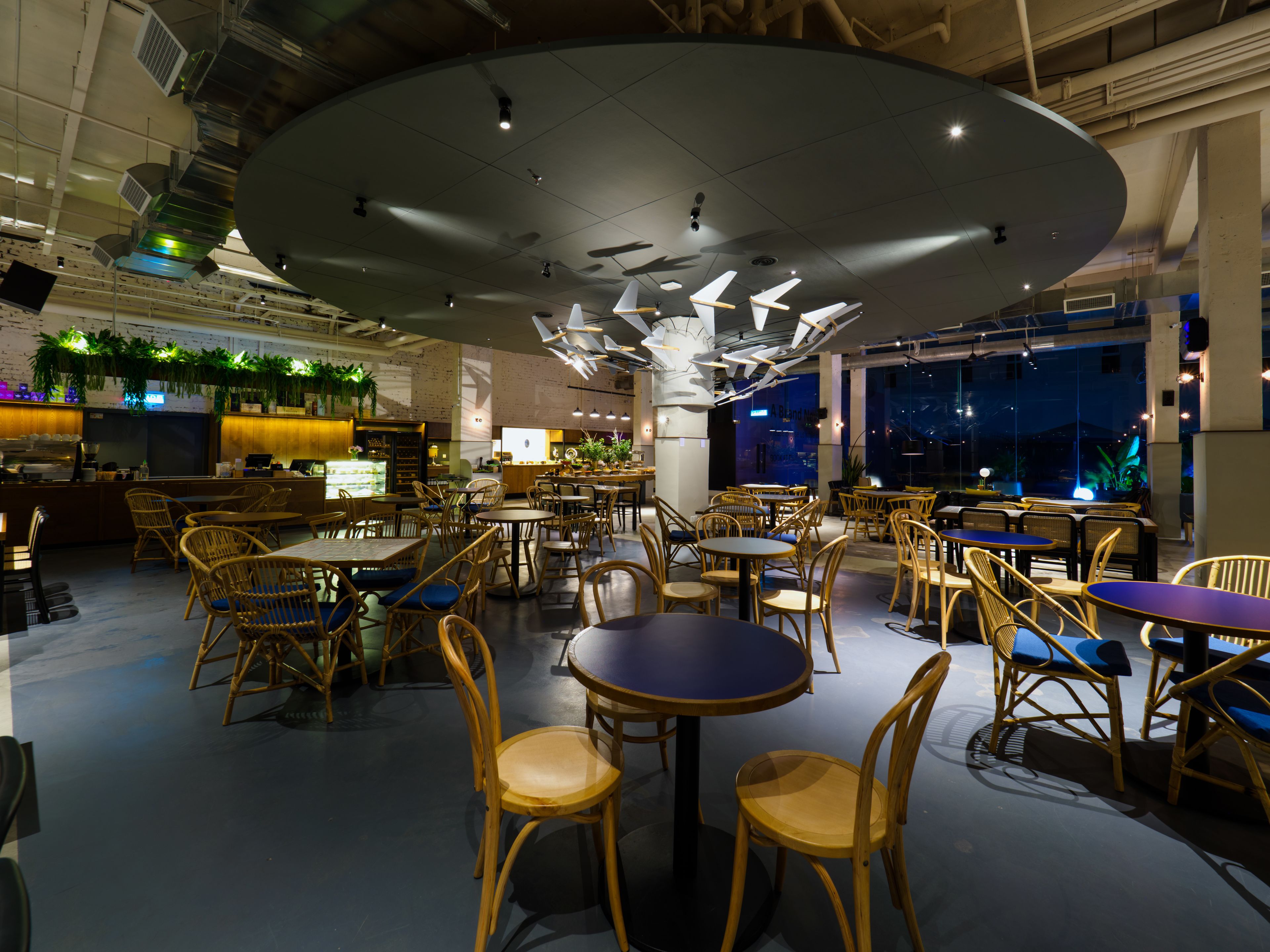 A spacious restaurant with high ceilings, modern lighting, and a variety of seating arrangements including round tables and wicker chairs.