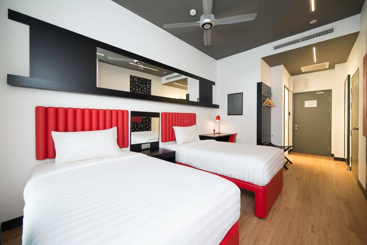 A hotel room with two beds, a red headboard, a large mirror, a ceiling fan, and a door leading to a hallway.