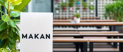 Letrero con la palabra 'MAKAN' en un ambiente moderno con bancos de madera y plantas en macetas.