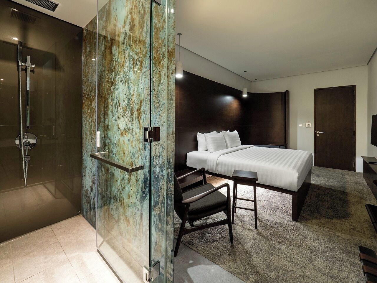Une chambre d’hôtel moderne dotée d’une douche à parois de verre, d’un lit avec du linge de lit blanc, d’une table de chevet en bois foncé et d’un petit bureau avec une chaise.