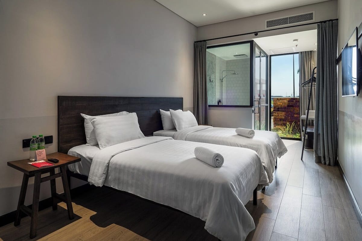 A hotel room with two beds, a small table with a bottle and a red object, and a view of a garden through a glass door.