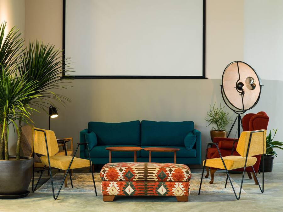 A living room with a teal sofa, two yellow chairs, a patterned ottoman, a floor lamp, and a large round mirror.